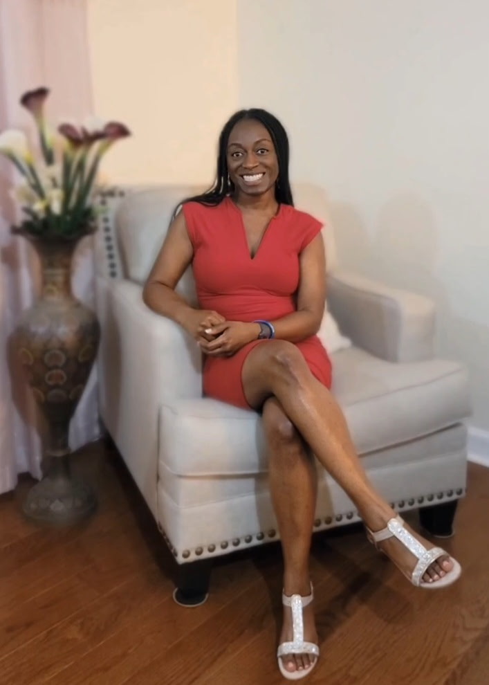 Portrait of DeLora Lambert seated in a cream chair, wearing a red dress and smiling warmly.